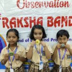 Three schoolchildren in beige uniforms pose with handmade badges in front of a banner saying observation of  at a school event each wearing a lanyard with id cards